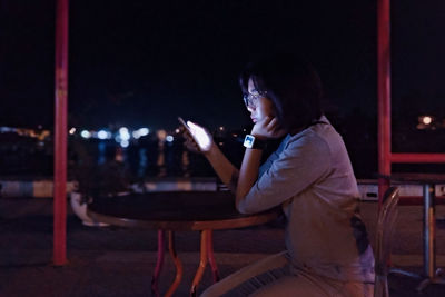 Side view of young woman using phone while sitting on chair at night