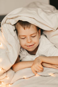 Portrait of a smiling boy lying under a white blanket and with a garland. child's christmas morning. 