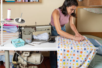 Professional hispanic seamstress making marks on textile at table with professional sewing machine and various supply during work in light workshop