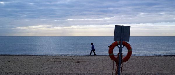 Panoramic view of woman walking at beach against cloudy sky during sunset