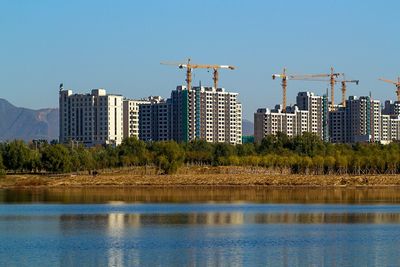 Cranes and buildings by lake against clear blue sky in city