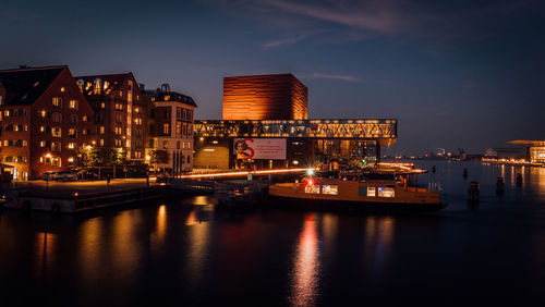 Illuminated buildings by river against sky at dusk