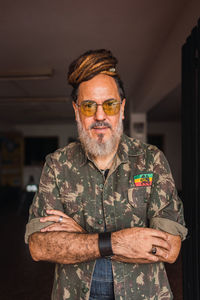 Bearded mature male in casual outfit with sunglasses and dreadlocks standing in light apartment and looking at camera
