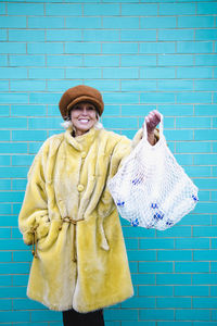 Smiling senior woman holding mesh bag with plastic bottles in front of turquoise brick wall