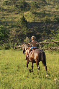 Rear view of shirtless boy riding horse on grassy field