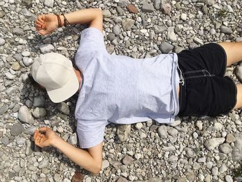 Man lying on rocky beach with baseball cap on his face