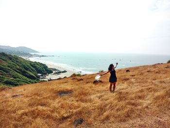 Woman with arms outstretched standing on grassy field against sea