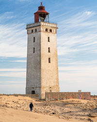 Rear view of woman walking on sand towards lighthouse at beach against sky