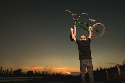 Rear view of young man with arms raised carrying bicycle while standing against sky during sunset