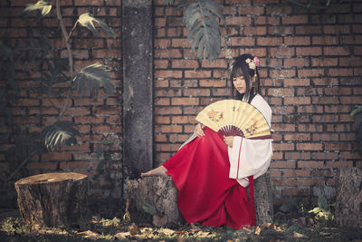 Young woman wearing traditional clothing while holding folding fan against brick wall