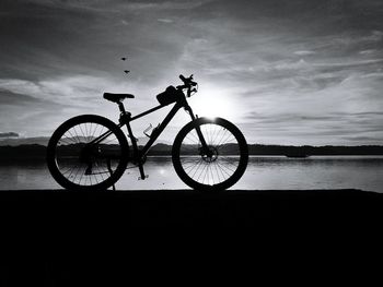 Silhouette bicycle by lake against sky during sunset