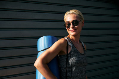 Sporty smiling blond woman in sportswear and sunglasses with yoga mat. workout outdoors. fitness