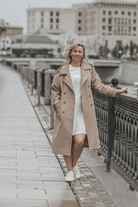 A middle-aged blonde woman with curly hair stands on the river embankment in the city.