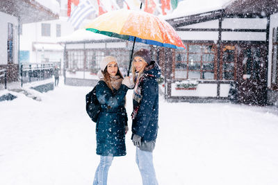 Two beautiful millennial caucasian women stand under a rainbow umbrella  under a snowfall.