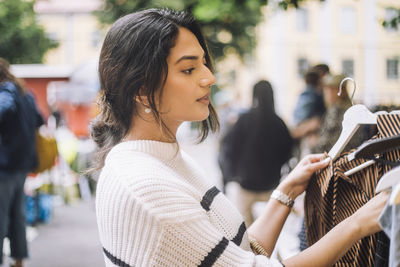 Side view of young female customer choosing dress while doing shopping during festival at flea market