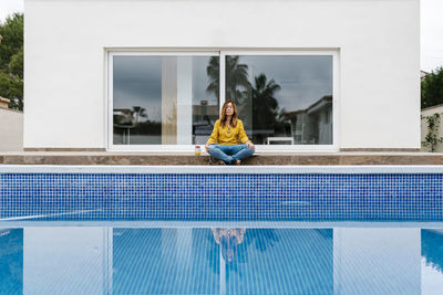 Mature woman meditating while sitting at poolside against house