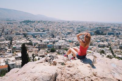 Rear view of woman sitting on rock formation while looking at city against sky