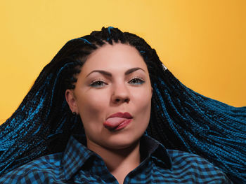 Portrait of young woman with braided hair sticking out tongue against yellow background