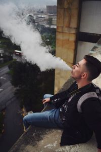 Man exhaling smoke while sitting on wall