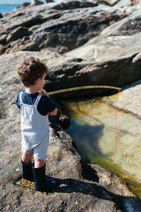 Side view of adorable child fisher in gumboots and overall holding small rod and fishing in shallow clear brook on rocks
