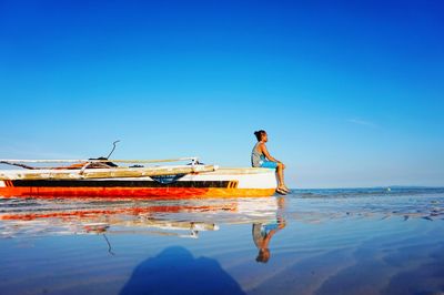 Side view of man sitting on boat moored at shore against blue sky