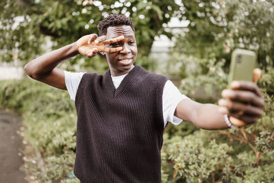 Young african man taking selfie on phone, show v sign near eye in park, smiling