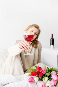 Woman sitting on the bed wearing pajamas, with pleasure enjoying flowers and a glass of red wine