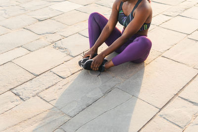 Crop black female in sportswear sitting on floor to do stretching exercises on a sunny day