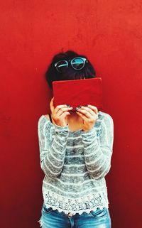 Woman holding book over face while standing against red wall at home