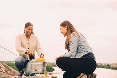 Smiling female friends roasting sweetcorns in picnic against sky