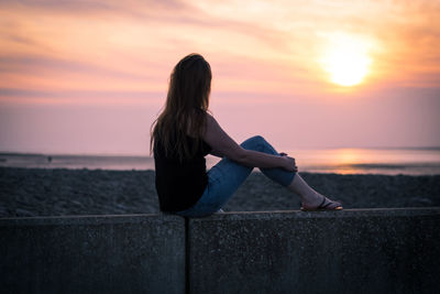 Woman sitting on retaining wall against sky during sunset