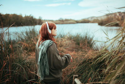 Side view of young woman wearing headphones while standing by lake against sky