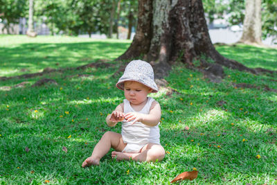 Adorable baby girl in white clothing and panama is playing with dry yellow leaves on grass.