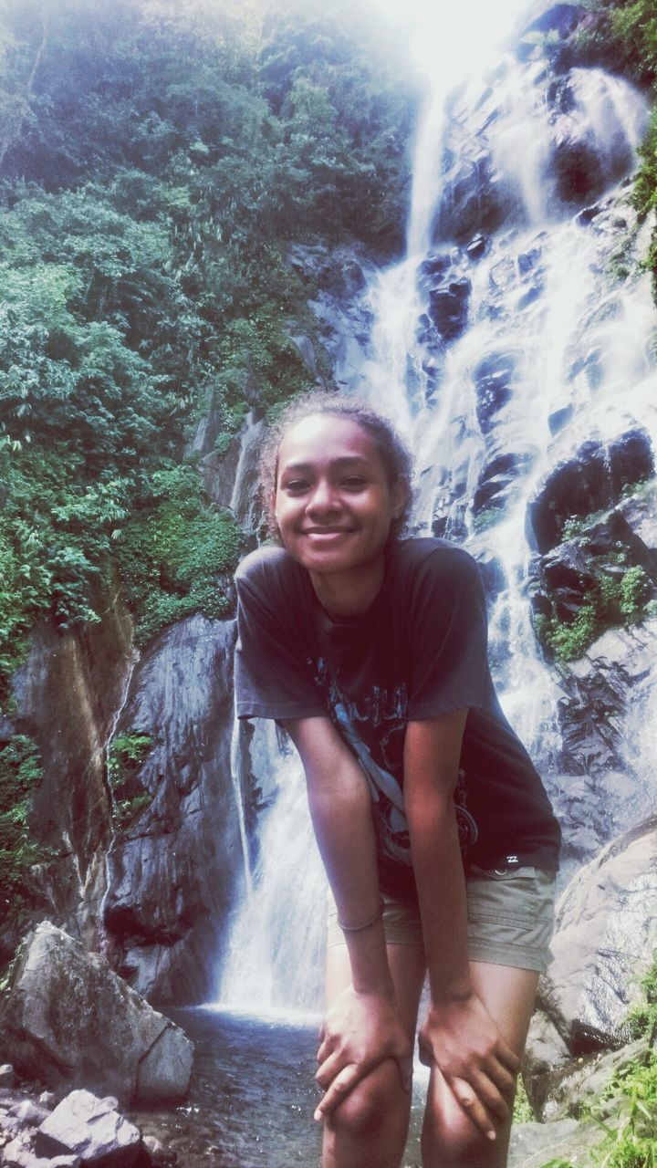 young adult, lifestyles, leisure activity, looking at camera, portrait, young women, person, water, tree, rock - object, front view, standing, waterfall, casual clothing, smiling, nature, three quarter length, forest