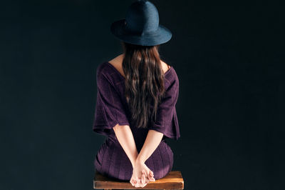 Rear view of young woman with hands clasped sitting on wooden stool against black background