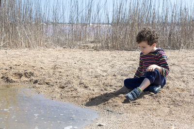 Small boy   with curly hair, looking with sand at the edge of pond on sunny day