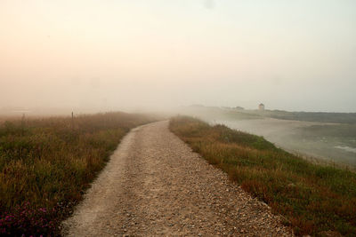 Road passing through landscape against sky during foggy weather