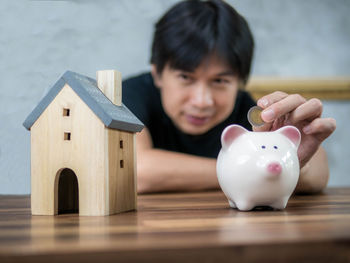 Portrait of man putting coin in piggy bank with model house on table