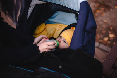 Mother feeding baby son from bottle in baby stroller outdoors in park