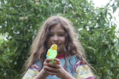 Low angle portrait of smiling girl holding toy gun against trees