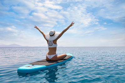 Rear view of woman with arms raised sitting on paddleboard in sea against sky