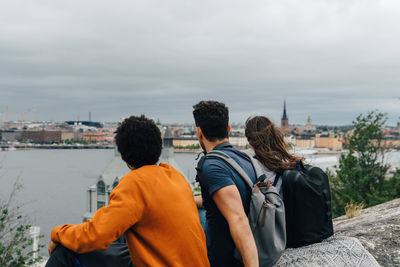 Rear view of friends sitting on hill by river in city against sky