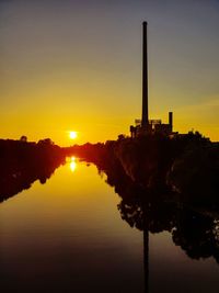 Silhouette factory by lake against sky during sunset