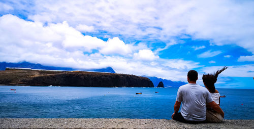 Rear view of couple sitting at beach against cloudy sky