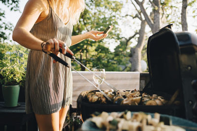 Midsection of woman grilling vegetables on barbecue while eating watermelon