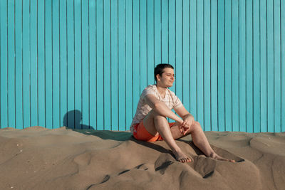 Young man looking away sitting in sand beach with turquoise wall