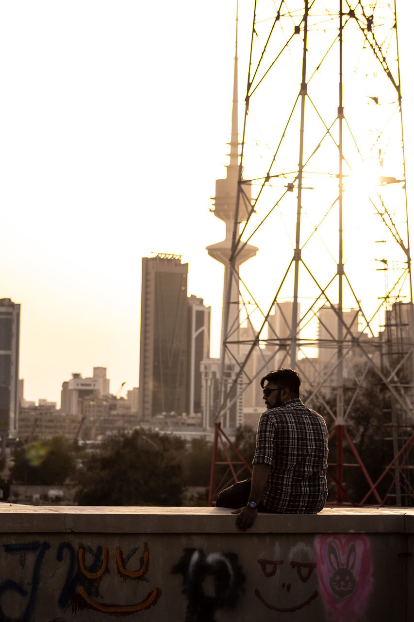 REAR VIEW OF MAN STANDING BY BUILDINGS IN CITY AGAINST CLEAR SKY