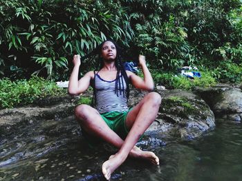 Young woman looking away while sitting on rock at lakeshore against trees