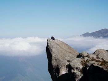 Couple sitting on top of cliff against clouds covering mountains