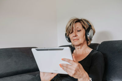 Senior woman sitting on couch, using on-ear headphones and tablet computer
